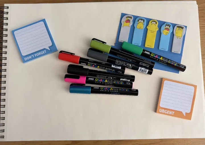 A3 sketchbook on a wooden desk with Osaka markers, monster stickers, and Don’t Forget and Urgent sticky notes